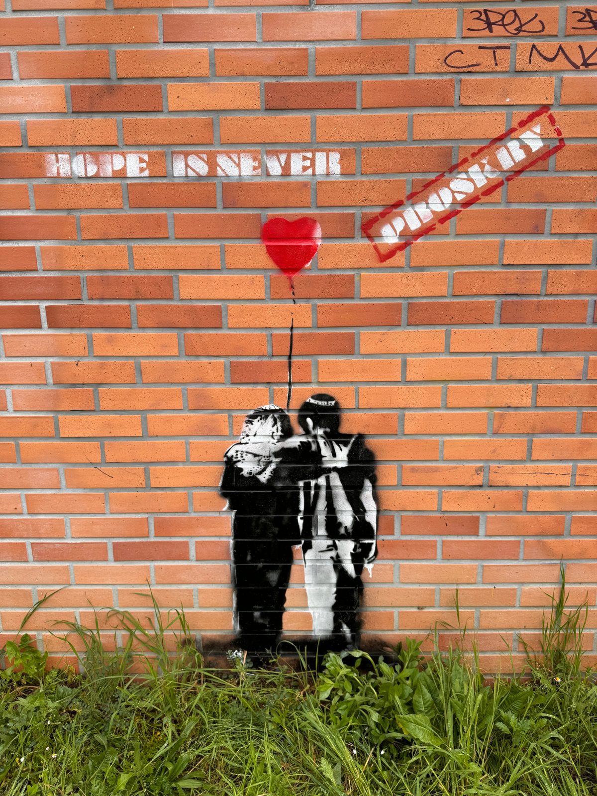 Pochoir Hope is never Proskry sur un mur de briques : la phrase au pochoir blanc, un ballon en forme de cœur rouge, le tampon Proskry en diagonale et un couple enlacé